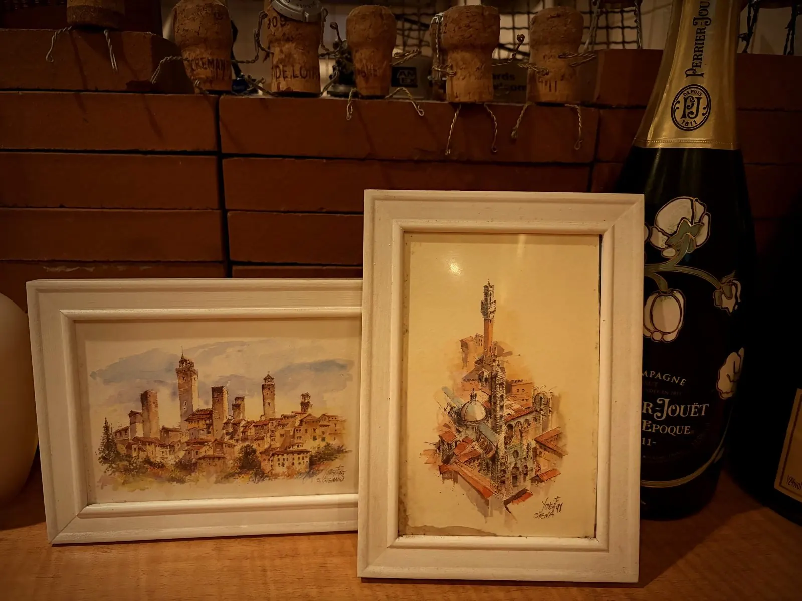 コルクが並ぶ棚の前に置かれた、イタリアの街を描いた小さな額2枚とシャンパーニュのボトル